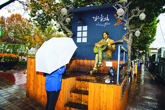 대구 방천시장 인근 ‘김광석 다시 그리기 길’을 찾은 탐방객이 우산을 쓴 채 기타를 치는 김광석 동상을 바라보고 있다. 이 길은 2008년 ‘문화를 통한 전통시장 활성화 시범사업(문전성시)’ 프로젝트의 하나로 추진됐다.
