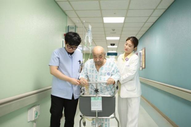 포괄병동에서 환자가 간호사와 보조원의 도움을 받아 복도를 이동하고 있다.