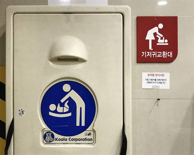 경기 고양시의 한 대형마트 남자 화장실에 부착된 두 개의 기저귀 교환대 안내표지가 대조적이다. 마트 측에서 부착한 안내표지(붉은색)는 치마 입은 여성이 주인공인 데 반해 교환대 제조사가 붙인 그림은 인물의 성 구분이 없다. 교환대 제조사는 외국 기업이다.