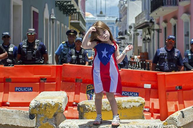 푸에르토리코 국기가 그려진 드레스를 입은 한 소녀가 18일 산후안 로세요 주지사 관저로 향하는 길을 막고 있는 경찰 앞에 서 있다. [AP=연합뉴스]