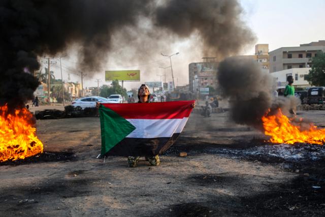 군부 쿠데타 반대 시위 참가자가 26일 타이어가 불타고 있는 수도 하르툼의 한 거리에서 수단 국기를 펼쳐들고 있다. 하르툼=EPA 연합뉴스