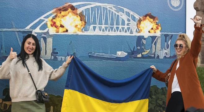 우크라이나 수도 키이우에서 지난 8일(현지시간) 대형 폭발사고로 붕괴된 크름대교(케르치해협 대교)의 모습이 그려진 벽화 앞에서 시민들이 우크라이나 국기를 들고 환호하며 기념사진을 찍고 있다. 키이우 | EPA연합뉴스