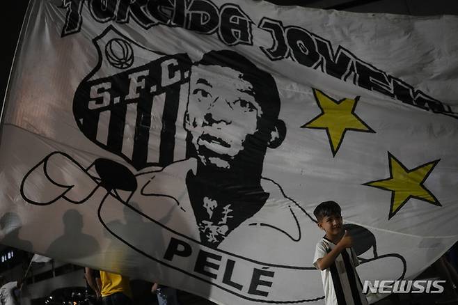 [산투스( 브라질)= AP/뉴시스] 펠레의 죽음이 보도된 29일(현지시간) 브라질 산투스 축구장 앞에서 휘날리는 펠레 초상 깃발 앞에서 한 소년이 엄지를 들어보이고 있다.