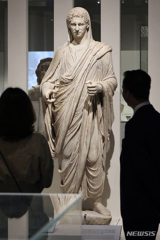 [서울=뉴시스] 박진희 기자 = '토가를 입은 남성의 초상' 대리석 전신상. 이 조각상의 주인공은 시민 중에서도 ‘제1의 시민’이라는 걸 드러내고 싶었던 로마 황제이거나 영향력이 강한 일반 시민이었을 것이다. 로마 남성 시민의 웃인 토가를 입었다는 것은 이 전신상의 주인공이 로마 시민권을 가진 자이고 노예나 이방인 같은 사회 집단과는 다르다는 점을 알려준다. 토가는 수 미터에 달하는 긴 모직 천을 셔츠 같은 튜닉 위에 걸쳐 입는 웃인데, 당대 유행에 따라 걸치는 방법이 달랐다. 이 작품에서도 볼 수 있듯이 로마 제정 시대에는 오른팔과 왼쪽 어깨 중간에 천 한 자락을 당겨 늘어지는 부분이 생기도록 하고 여기에 물건을 담아 주머니처럼 사용했다. 토가의 색이나 형태는 사회적 지위를 나타내는 역할을 하기도 했다. 예를 들어 원로원은 보라색 줄무늬가 있는 토가 를 입을 수 있었고, 성공적인 원정을 기념하는 개선 행렬에서 황제는 값이 무척 비싼 보라색 염료로 전체를 물들인 토가를 입었다. 2023.06.17. pak7130@newsis.com