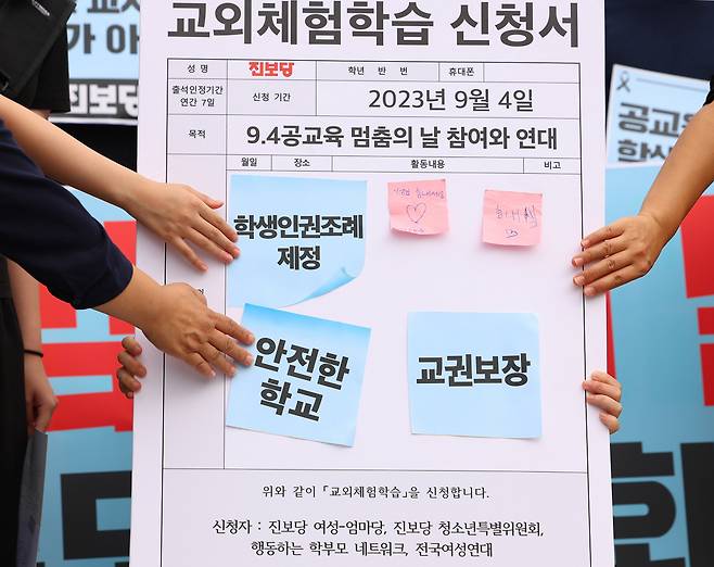 서울 서초구 초등학교에서 숨진 교사의 49재이자 이를 추모하는 '공교육 멈춤의 날'인 4일 오전 서울 광화문광장 이순신 장군 동상 앞에서 진보당 여성-엄마당 관계자 및 학부모·학생이 '공교육 멈춤의 날' 지지 기자회견을 마친 뒤 현장체험학습 판에 표어를 붙이는 퍼포먼스를 하고 있다. 이날 기자회견에는 당일 현장체험학습을 낸 가족과 청소년, 학부모가 함께 참가했다. [연합]