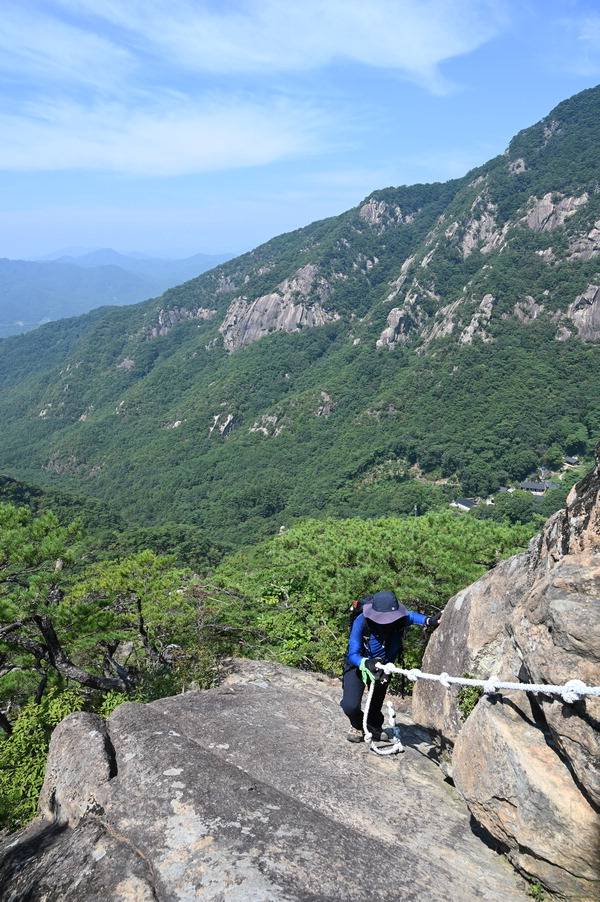 남근석을 지나 로프가 묶인 암릉을 오르는 취재팀 뒤로 배바위와 무암사가 보인다.