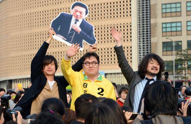 2012년 4월 8일 민주통합당(더불어민주당 전신) 총선 후보로 나선 김용민(노원갑) 후보가 서울광장에서 열린 인터넷 팟캐스트 '나는꼼수다' 팬 미팅에서 참석자들의 환호에 화답하고 있다. 나꼼수 멤버인 주진우(왼쪽) 시사인 전 기자와 김어준씨가 또 다른 멤버인 정봉주 전 의원의 사진을 들고 함께 서 있다. 한국일보 자료사진