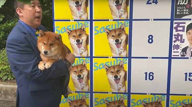 일본 정치 단체 'NHK로부터 국민을 지키는 당' 다치바나 다카시가 자신의 반려견을 들고, 반려견 사진으로 도배된 도쿄도지사 선거 입후보자 게시판을 소개하고 있다./유튜브