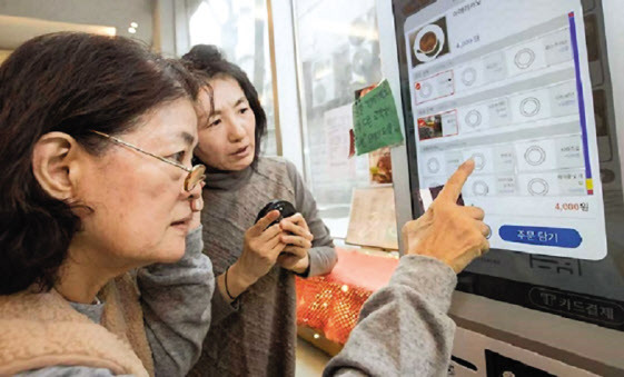 한 노인이 키오스크 이용에 어려움을 겪다 주변 사람의 도움을 받아 커피를 주문하고 있다.