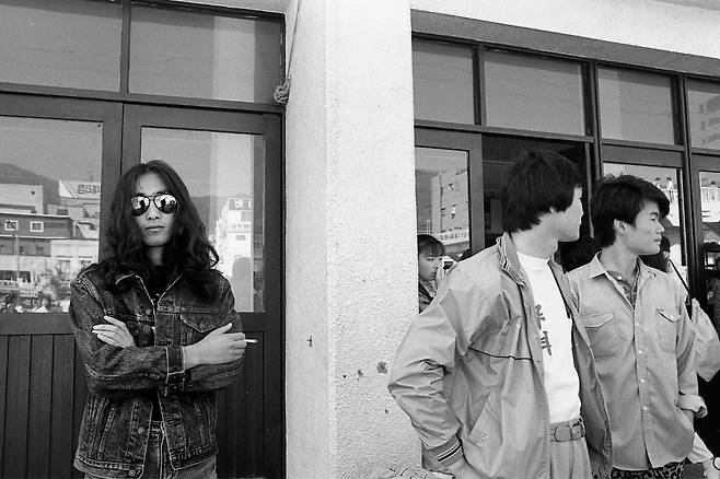 1987년 부산진역 앞에서 선글라스를 끼고 청자켓을 입은 한 청년이 카메라를 보고 있다. 오른쪽에 '무역'이라고 쓴 과티(과티셔츠)를 입은 대학생이 보인다.당시 부산진역은 비둘기호 같은 완행열차의 출발역으로 대학생들이 엠티를 가기 위한 집결지로 유명했다. /사진가 문진우