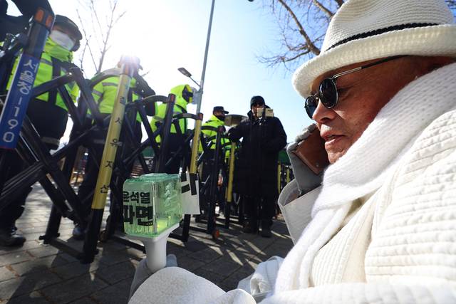 ▲ 지난해 12월 26일 오후 서울의소리 백은종 대표가 서울 용산구 한남동 대통령 관저 인근에서 경찰들이 이동을 제한하자 바리케이드 앞에 앉아 통화하고 있다. 연합뉴스
