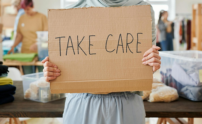 TAKE CARE이라고 쓰여있는 종이를 손에 들고 병원에 서있는 원피스를 입은 사람