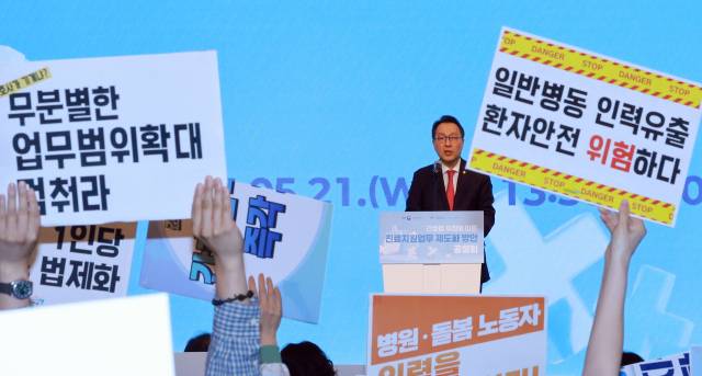 박민수 보건복지부 제2차관이 21일 서울 용산구 피크앤파크 컨벤션 로얄홀에서 열린 간호법 제정에 따른 진료지원업무 제도화 방안 공청회에 참석해 개회사하는 가운데 전국민주노동조합총연맹 공공운수노조 의료연대본부 조합원이 피케팅하고 있다.연합뉴스