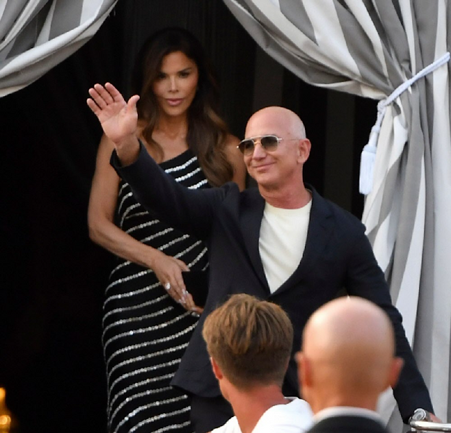 25일(현지 시간) 이탈리아 베네치아의 한 호텔에서 제프 베이조스 아마존 창업자(오른쪽)가 손을 흔들어 보이고 있다. 뒤에 선 여성은 그의 약혼자 로런 산체스. 둘은 26일부터 사흘간 베네치아에서 초호화 결혼식을 연다. 베네치아=AP 뉴시스