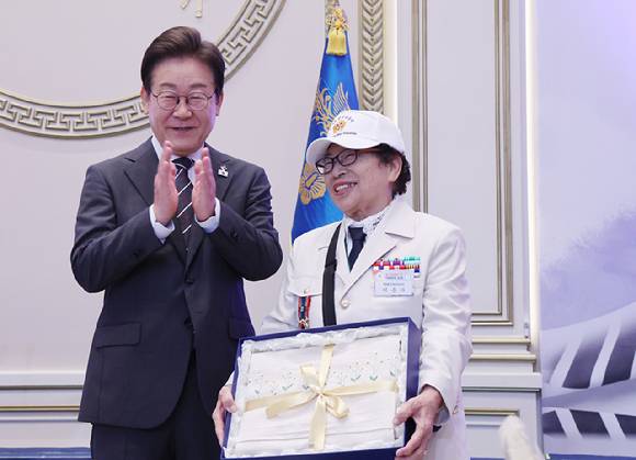 이재명 대통령이 27일 청와대 영빈관에서 열린 '호국 보훈의 달, 대통령의 초대' 행사에서 특별 초청대상자인 이춘자 씨에게 광목이불을 선물한 뒤 박수치고 있다. [사진=연합뉴스]