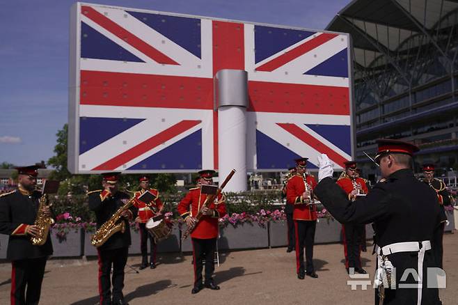 [애스콧( 영국)=AP/뉴시스]  2025년6월 18일 영국 애스콧 왕립 경마대회장에서 거대한 전광판 국기 앞에서 연주하는 군악대.  영국왕실은 왕실 비용을 절감하고 국가적 현대화에 부응하기 위해 왕실전용 열차 운행을 폐지하기로 했다고 6월 30일 발표했다. 2025. 07.01.