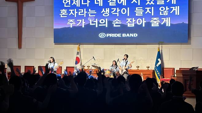 프라이드밴드가 교회에서 찬양 사역을 하는 모습. 스크린에는 "언제나 네 곁에 서 있을게 혼자라는 생각이 들지 않게 주가 너의 손 잡아 줄게"라는 GOD '촛불하나'를 개사한 가사가 떠 있다. DSM엔터테인먼트 제공