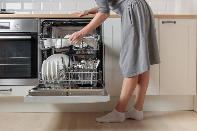 하얀색 주방에서 붙박이 식기세척기를 여는 여성. 내부에는 세척이 완료된 접시와 식기가 가지런히 놓여 있다. 시간 절약형 주방가전의 한 장면. 출처=123rf