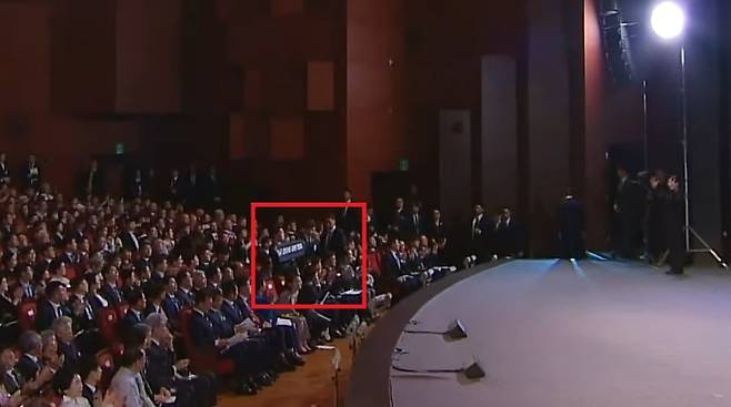 광복절 경축식 무대 전경 내, 관객석 중앙에 안철수 의원이 ‘조국·윤미향 사면 반대’ 펼침막을 들고 있는 모습이 포착됐다. 대통령이 무대에서 퇴장하는 동선에 위치해 해당 장면이 행사장 중앙 시야에 그대로 드러났다. (유튜브 캡처)