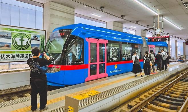 타카오카시역 트램 정류장에 도라에몽 트램이 들어서자 관광객이 사진을 찍고 있다.