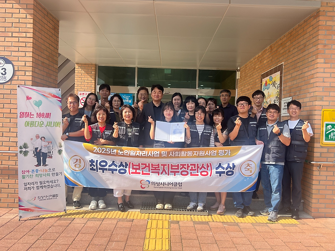 ▲ 29일 오후 경북 의성군 의성시니어클럽 청사 앞에서 김성진 관장(뒷줄 중앙)과 직원들이 보건복지부 장관상(앞줄 중앙)을 들고 현수막을 펼친 채 손가락 하트를 하며 2년 연속 최우수 수행기관 선정을 축하하고 있다. 의성시니어클럽