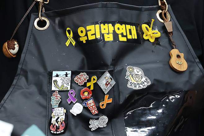 주휘씨가 메고 있는 앞치마에는 우리밥연대가 다닌 다양한 투쟁 현장들의 배지가 달려있다. 거꾸로 뒤집어쓰는 캡모자에는 세월호 참사를 상징하는 노란 리본 배지가, 손목에는 노란색(세월호 참사)·보라색(이태원 참사)·하늘색(아리셀 참사) 팔찌가 있다.