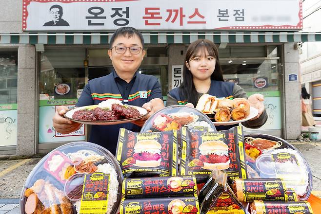 서울 관악구에 있는 매운맛 돈가스 식당 ‘온정돈까스’ 본점 앞에서 송중원 온정돈까스 대표(왼쪽)와 세븐일레븐 직원이 협업 상품들을 선보이고 있다. 세븐일레븐 제공