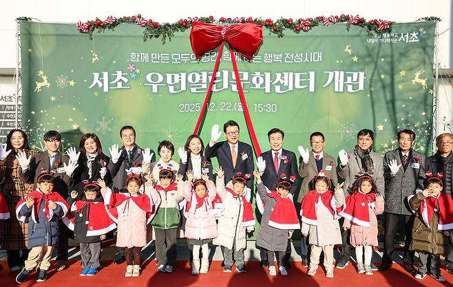 단체 사진으로, 뒷 배경에는 덩굴 장식과 리본으로 장식된 ‘서초 우면열린문화센터 개관, 2025년 2월 22일 15시 30분’이라고 쓰여 있는 현수막이 걸려 있다. 정장 차림의 성인들과 망토를 입은 아이들이 함께 손을 들고 있다.