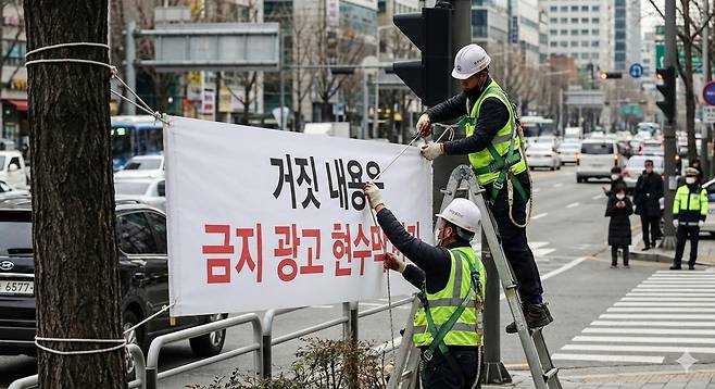 거짓 내용을 금지 광고 현수막에 게시한 현수막을 제거하기 위해 안전 장비를 착용한 작업자들이 사다리 위에서 작업하고 있는 사진이다. 현수막에는 ‘거짓 내용은 금지 광고 현수막’이라는 텍스트가 적혀 있다.