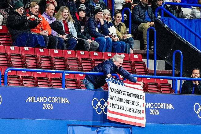 밀라노에서 피겨 스케이트 경기를 관람하던 한 관중이 미국 국기 위에 “세계에 사과드립니다. 우리 스스로 고칠 거에요!”라고 쓴 사과 펼침막을 들어 보이고 있다. 뉴욕타임스 8일 보도 갈무리.