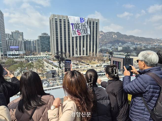 방탄소년단(BTS) 컴백 공연을 하루 앞둔 20일 서울 종로구 대한민국역사박물관 옥상정원에서 관광객들이 맞은편 정부서울청사 건물에 설치중인 대형 현수막을 촬영하고 있다. 김태욱 기자