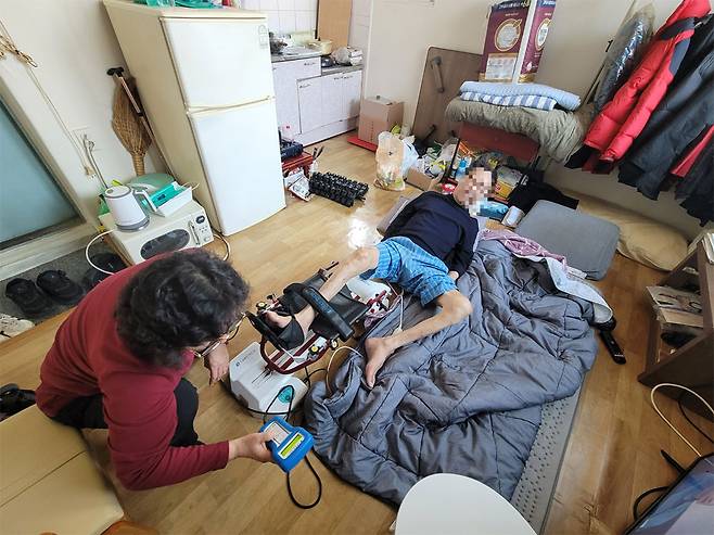 ▲ 김상훈(68·가명) 씨가 요양보호사의 도움을 받아 다리 재활 기기를 사용하고 있다. 지난 2024년 10월 고관절 골절을 당한 김 씨는 거동이 어려워 누워 생활한다.