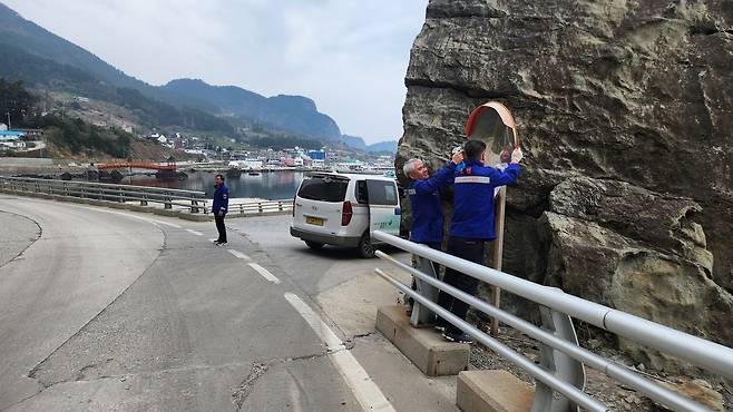 거대한 바위 절벽을 끼고도는 울릉도 해안 일주도로의 굽은 길목에서 가드레일 밖 좁은 발판 위에 올라서 곡면거울(반사경)의 시야를 가리는 오염물을 닦아내고 있다. /모범운전자회 울릉지회 제공
