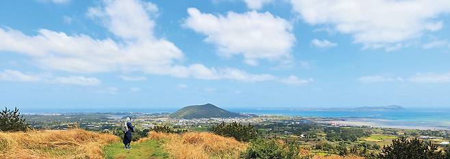 제주 올레길 1코스의 하이라이트 구간인 알오름 정상을 한 여행자가 걷고 있다. 탁 트인 하늘과 다양한 채도의 제주 바다, 그리고 밭담을 두른 마을을 내려다보며 걷는 길이다. 멀리 야트막한 산처럼 보이는 곳이 제주 올레길 마지막 21코스가 지나는 오름 지미봉이다.