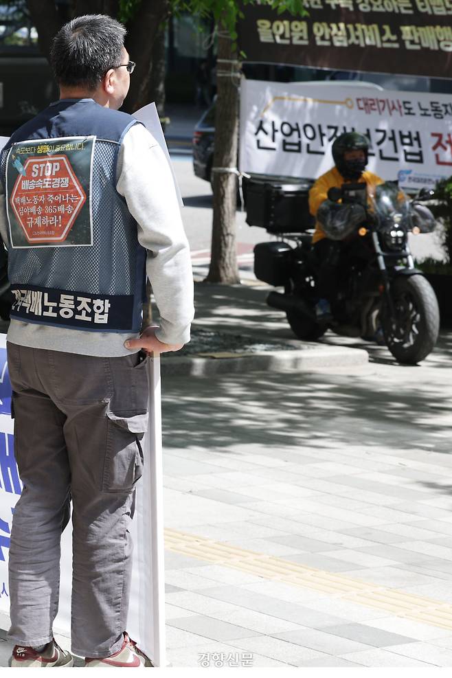 택배노조가 ‘택배사의 노동절 휴업일 미지정 규탄 및 특수고용노동자 차별 철폐 기자회견’을 하고 있는 가운데 한 배달 노동자가 앞을 지나가고 있다. 운송 노동자 대부분은 노동절 법적 휴무 대상자가 아니다.