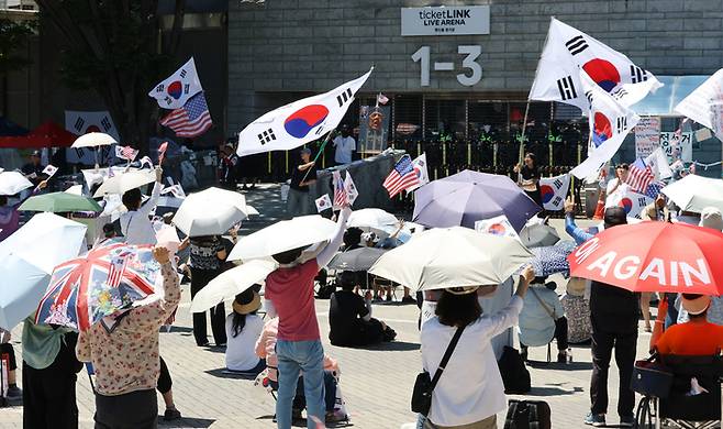 낮 최고 기온이 29도로 예보된 지난 23일 6·3 지방선거 잠실 개표소로 사용된 서울 송파구 올림픽공원 핸드볼경기장 앞에서 투표용지 부족 사태 규탄 봉쇄 시위 참가자들이 우산을 쓰고 구호를 외치고 있다. [연합뉴스]