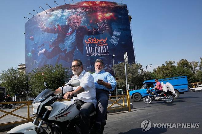 2026년 8월 24일 이란 테헤란에서 오토바이 운전자들이 폭발 속에서 물에 빠진 도널드 트럼프 미국 대통령의 모습과 함께 "위대한 승리! 호르무즈 해협"이라는 문구가 적힌 대형 광고판 앞을 지나가고 있다. [AP=연합뉴스 자료사진. 재판매 및 DB 금지]