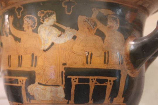 러시아 상트페테르부르크 예르미타시 박물관에 보관된 BC 4세기 그리스 도자기. 심포지온에 참석한 3명의 남자가 긴 의자에 앉아 포도주를 마시는 동안 헤타이라가 아울로스를 불며 흥을 돋운다.