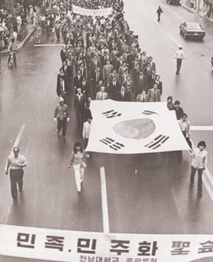 대형태극기를 앞세우고 민족민주화대성회 참석을 위해 교문을 벗어나 금남로로 향하고 있는 전남대학교 교수들, 이들 뒤를 학생들이 따르며 민주주의를 위한 구호를 외치고 있다. ⓒ5·18기념재단