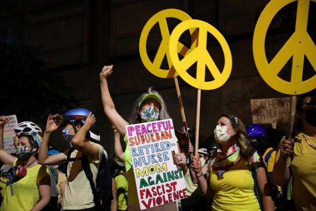 포틀랜드에서 21일 노란 옷을 입은 엄마들이 인종차별 반대 및 연방요원 파견에 항의하며 시위를 하고 있는 가운데 한 시위자가 '평화로운 교외의 은퇴한 간호사 엄마, 파시스트 폭력 반대'라고 적힌 손팻말을 들고 서 있다. 포틀랜드=로이터 연합뉴스