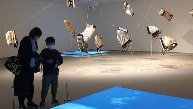 인천 영종도 파라다이스 아트스페이스에서 관람객이 허먼 콜겐의 작품 ‘어반 윈드’를 살펴보고 있다. 전시장 바닥의 영상은 캐나다 몬트리올 하늘을 보여주고, 공중에 매달려있는 아코디언들은 영종도 바람 데이터를 전기신호로 받아서 음악 소리를 낸다.