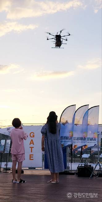 ▲ 지난 7월 28일 망상해수욕장 북쪽 끝자락에 위치한 망상오토캠핑리조트 앞 해변 데크에 마련된 ‘드론배송존’애서 디스이즈엔지니어링이 운용하는 ‘드론음식배송서비스’에 치킨을 주문한 고객이 치킨을 싣고 70m 상공위 1.5km의 하늘길을 비행해 배송존 위에 도착, 착륙하고 있는 드론을 사진을 찍으며 신기한 듯 바라보고 있다.
