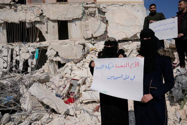 지난 2월 시리아 아타렙에서 지진 피해 주민들이 '유엔은 시리아인들을 죽이는 바샤르 아사드 대통령과 한통속'이라고 쓰인 손팻말을 들고 파괴된 집 잔해에 서서 시위하고 있다. 아타렙=AP 뉴시스
