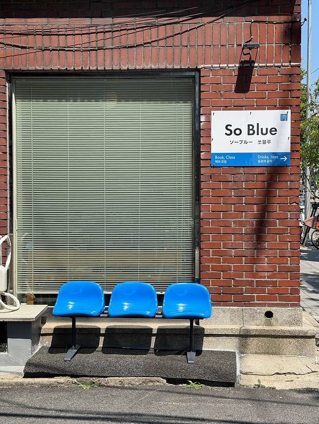 붉은 벽돌 건물 외벽에 걸린 '쏘블루(So Blue)'의 표지판. '책과 모임(Book, Class)'과 '음료와 음악(Drinks, Jazz)'이라는 문구 아래, 선명한 파란색 좌석 세 개를 놓았다.(사진=신문경 기자)